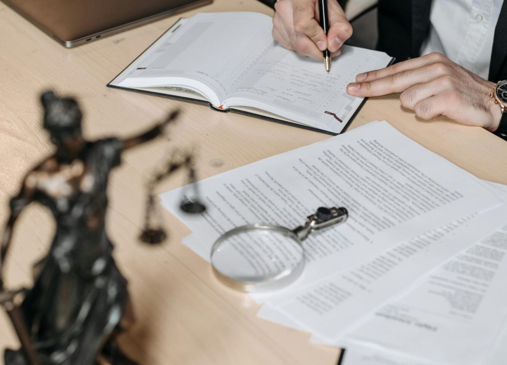 Professional working on documents at desk with legal statue and magnifying glass.